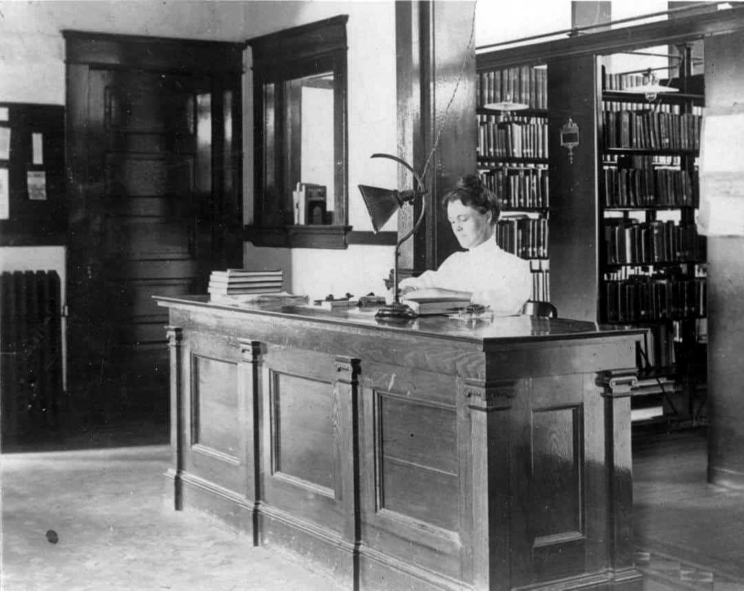 Mary Cornelia Lee is sitting behind a wooden desk stamping a book.